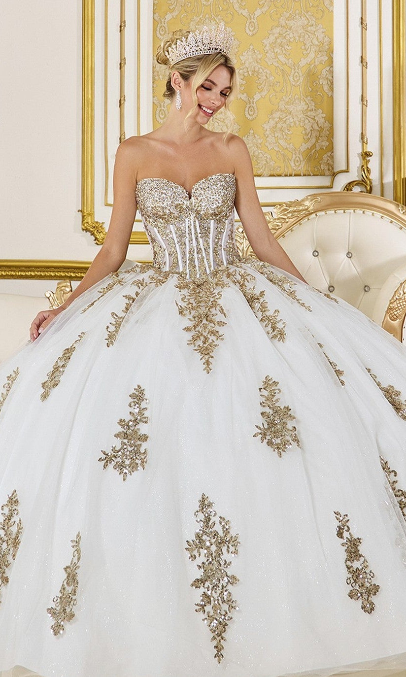 A woman in the Quinceanera Dress 5085 By Dancing Queen, featuring an ivory strapless sweetheart corset with gold sequin applique, stands in an ornate gold-accented room, crowned with a jeweled tiara.